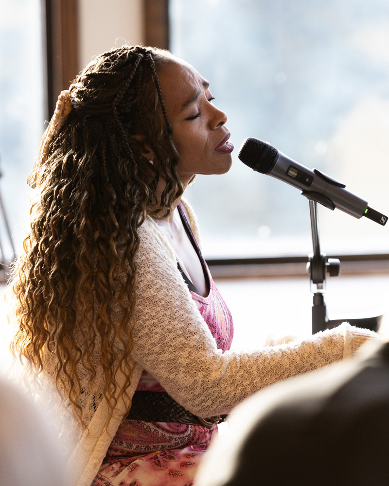 E'lissa Jones singing into a handheld microphone, eyes closed in a performance setting.