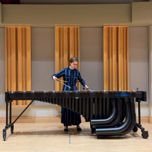 Person wearing a dark blue outfit playing a large black marimba with mallets on a stage with wooden wall panels behind.