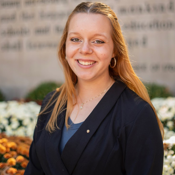 Smiling young woman with long blonde hair in a navy blazer and blue top, standing outdoors near flowers and a blurred wall backdrop.