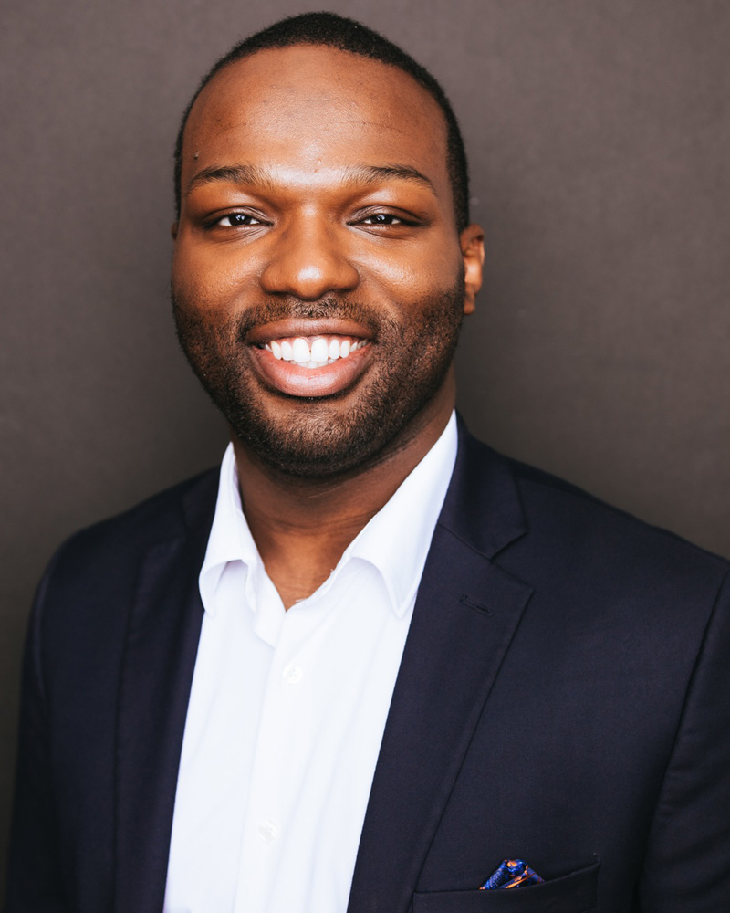 Shavon Lloyd in a navy blazer and white shirt, head-and-shoulders portrait against a dark backdrop.