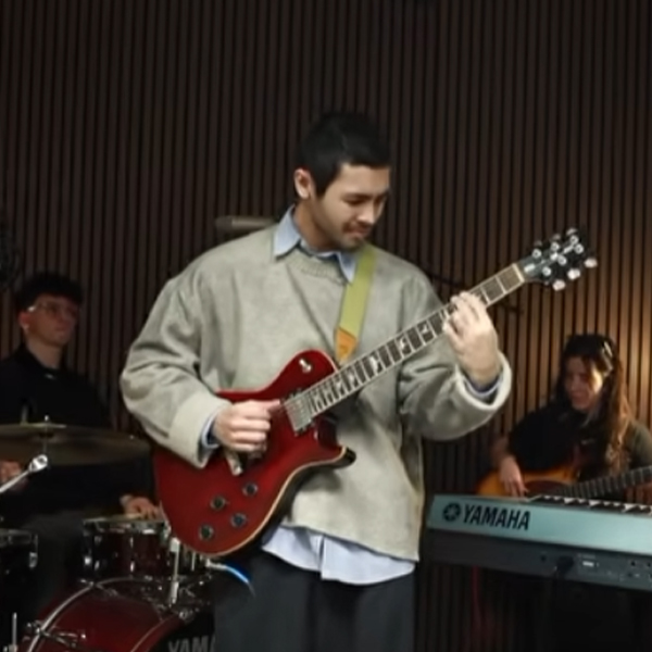 Guitarist in a gray sweater performs on stage, playing a red electric guitar while bandmates accompany on drums and keyboard.